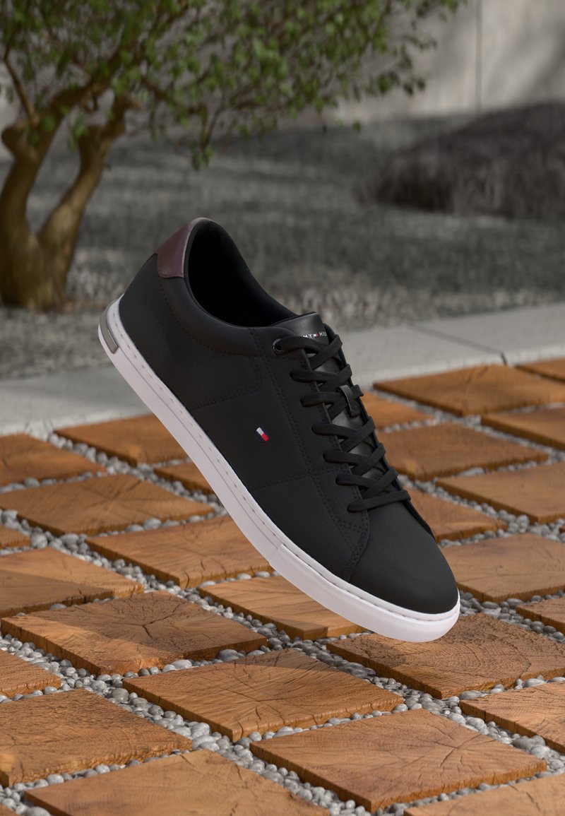 Zwarte sneakers met een glad synthetisch bovenwerk, witte rubberen zool en kastanjebruine hielaccent. Voorzien van zwarte veters en een klein logo detail.