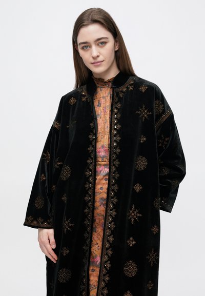 Jauna moteris su ilgais rudais plaukais, dėvinti juodą aksominį paltą su auksinėmis siuvinėjimo detalėmis virš gėlėto raštuoto suknelės, stovinti priešais paprastą foną.