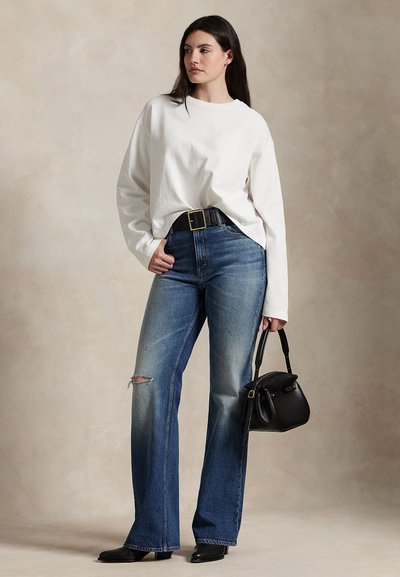 Kvinde står, iført hvid langærmet skjorte, blå højttaljede jeans med hul på venstre knæ, sort bælte, sorte støvler, holder sort håndtaske.