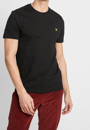 Man draagt een nauwsluitend zwart T-shirt met korte mouwen en een klein geel vogel-logo op de borst en rode corduroy broeken.