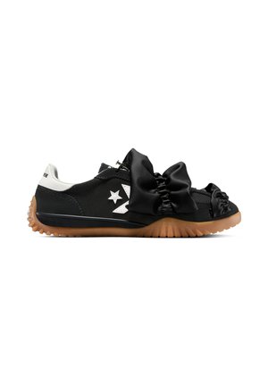 Zapatilla negra de perfil bajo con logo blanco, detalles de tela negra fruncida en la parte superior, lengüeta blanca en el talón y suela estriada color goma.