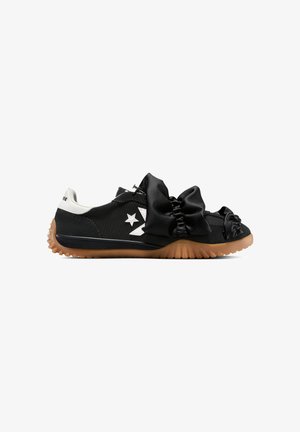 Zapatilla negra de perfil bajo con logo blanco, detalles de tela negra fruncida en la parte superior, lengüeta blanca en el talón y suela estriada color goma.