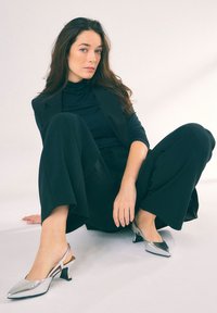 Srebrne czółenka na obcasie z odkrytym piętą, o spiczastych noskach i niskim słupku, zestawione z czarnym golfem i spodniami z szerokimi nogawkami na neutralnym tle.