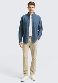 Blauwe gebutton-down shirt van gestructureerde stof, gedragen over een witte T-shirt; gecombineerd met beige broeken en witte sneakers.