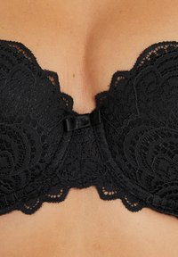 Reggiseno in pizzo nero con bordi ondulati, motivi floreali e un piccolo fiocco di raso al centro. Texture liscia con supporto a balze.
