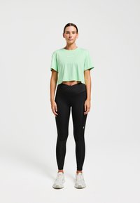 T-shirt court vert clair à manches courtes, associé à des leggings noirs taille haute. Baskets blanches et logo minimaliste sur le t-shirt.