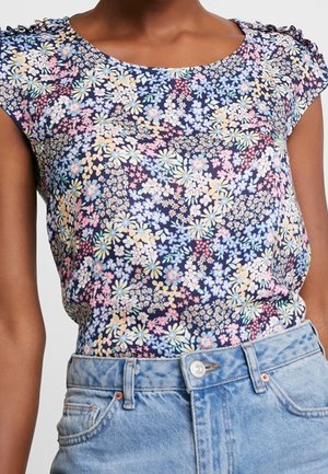 Kvinde iført en blomstret kortærmet bluse med blå, pink og gule blomster, stoppet ind i højtaljede lyseblå cowboybukser.