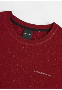 Burgundowa koszulka z okrągłym dekoltem wykonana z miękkiego materiału, z plecionym wzorem i małą wytłoczoną metką z logo z przodu.