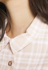 Košile s límečkem v béžovém károvaném vzoru s bílými linkami, vyrobená z hladké látky. Obsahuje velké středové knoflíky.