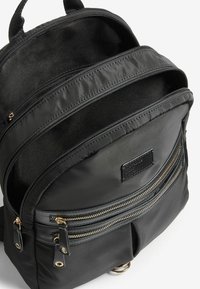 Sac à dos en nylon noir avec plusieurs compartiments, doté de zips dorés, d'une surface texturée et d'une étiquette sur la poche avant.