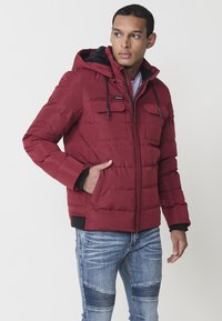 Crvena puffer jakna s kapuljačom, koja ima dva džepa na prsima i prednji patentni zatvarač. Materijal izgleda glatko s mekim podstavom u kapuljači.