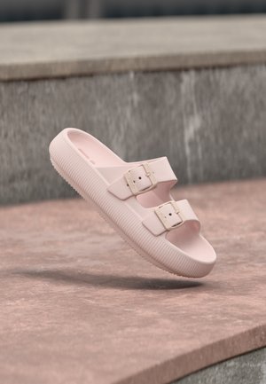 Lyserød slip-on sandal med to justerbare remme med spænder, tyk tekstureret sål, placeret på en stenovertflade nær en væg.