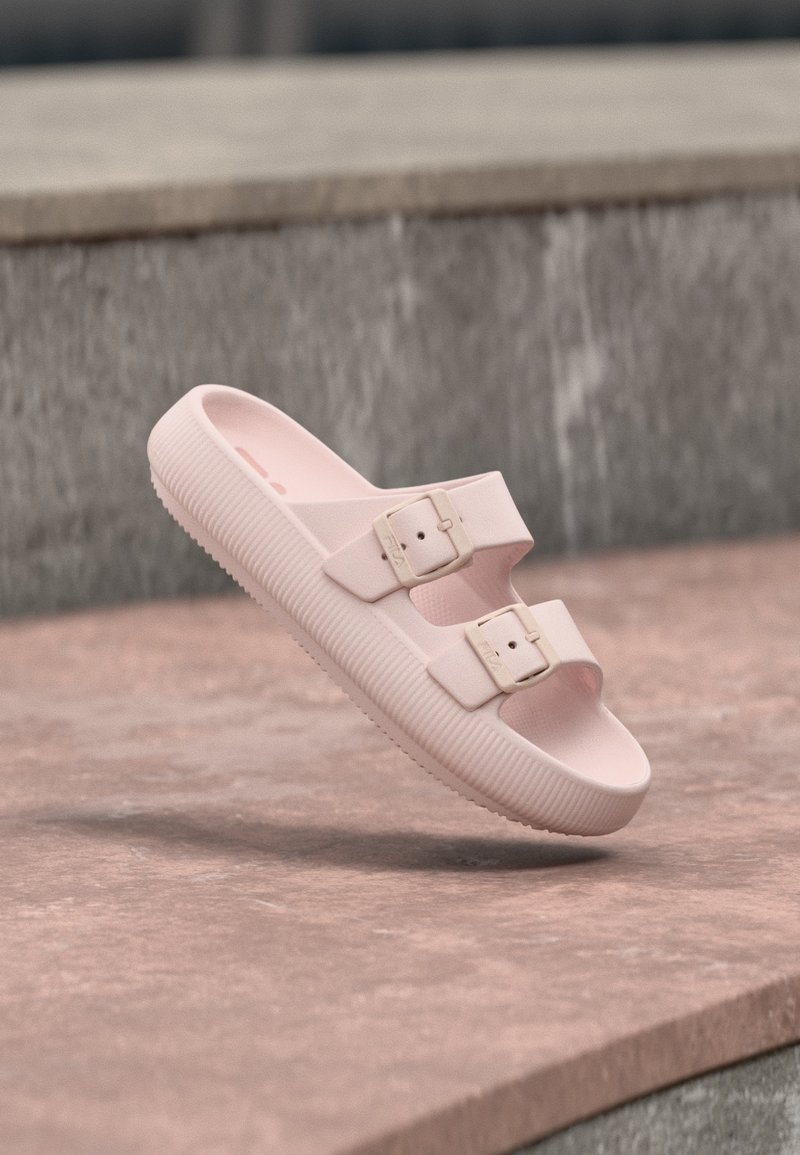 Ροζ ανοιχτό σανδάλι με δύο ρυθμιζόμενους ιμάντες με αγκράφες, χοντρή υφασμάτινη σόλα, τοποθετημένο σε πέτρινη επιφάνεια κοντά σε έναν τοίχο.