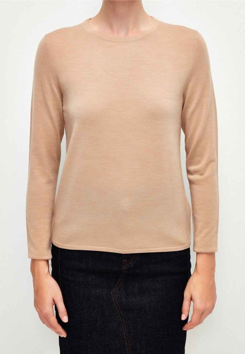 Femme portant un haut à manches longues beige et une jupe en denim foncé, debout devant un fond blanc uni.