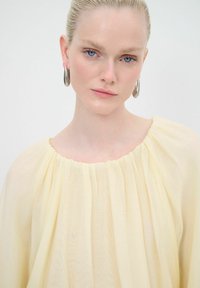 Helles gelbes Blusenshirt mit weicher, fließender Textur und gerafftem Ausschnitt. Das Model trägt silberne Creolen. Einfaches, elegantes Design.