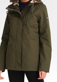 Veste imperméable vert olive avec zip sur le devant, capuche détachable et doublure douce. Comprend des poches et des accents de logo. Texture lisse.