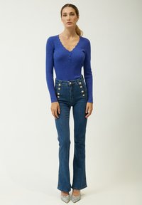 Top lungo a maniche corte blu con scollatura a onde, abbinato a pantaloni in denim blu a vita alta con fondo svasato e dettagli in bottone dorato.