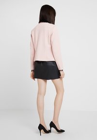 Veste rose texturée à coupe ajustée, associée à une jupe noire. Le modèle porte des escarpins noirs à talons aiguilles.