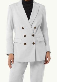 Blazer blanco de doble botonadura con tejido texturizado y botones marrones. Presenta solapas con muesca y bolsillos laterales. Combinado con pantalones a juego.