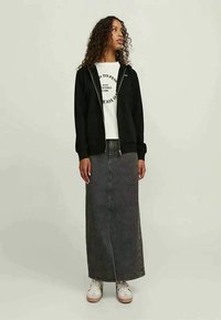 Donna con capelli ricci che indossa una felpa nera con zip, una maglietta bianca con grafica, una lunga gonna di jeans scuri e sneakers bianche, in piedi su uno sfondo semplice.