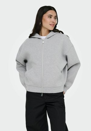 Donna con capelli lunghi e scuri che indossa una felpa grigia con zip e pantaloni neri, in piedi con le mani nelle tasche, guarda di lato.