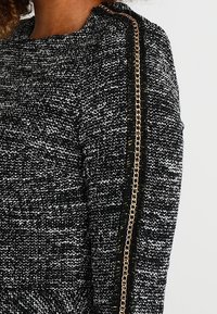 Maglione testurizzato bianco e nero con dettaglio a catena metallica dorata lungo la cucitura della spalla. Il tessuto ha un aspetto intrecciato.