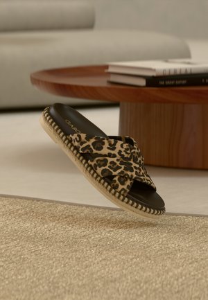 Sandale à glissière imprimé léopard avec semelle espadrille tissée flottant au-dessus d'un tapis beige tissé près d'une table ronde en bois.