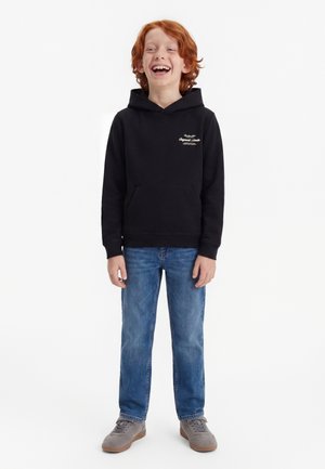Enfant roux riant, portant un sweat à capuche noir, un jean bleu et des baskets grises, debout sur un fond blanc.