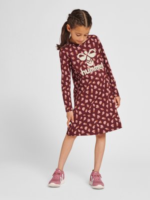 Dziewczynka w bordowej sukience z długim rękawem z różowym wzorem serc i logo „hummel”, ubrane w różowe trampki, stoi z opuszczoną głową.