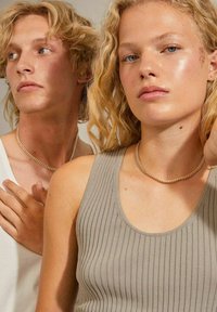 Zwei Models mit welligem Haar tragen schlichte, figurbetonte, gerippte Tops; eines in Grau, das andere in Weiß. Beide haben metallische Halsketten und neutrale Hintergründe.