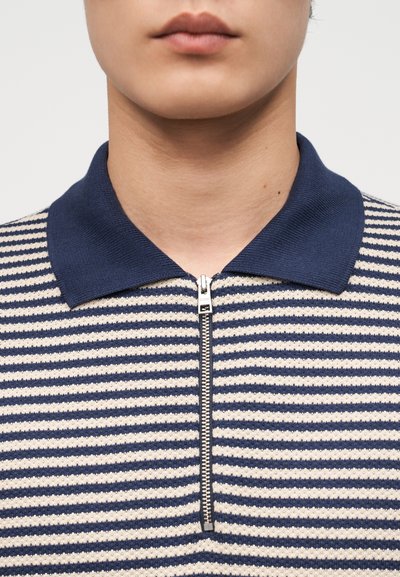 Κλείσιμο σε πρώτο πλάνο ενός ατόμου που φοράει μπλε ναυτικό και λευκό ριγέ πόλο με κοντό φερμουάρ στο μπροστινό μέρος και γιακά σε μπλε ναυτικό χρώμα.