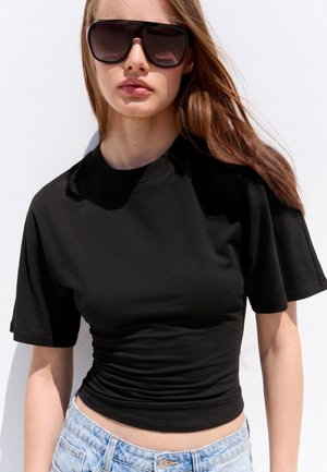 Jeune femme aux longs cheveux portant de grandes lunettes de soleil noires, un t-shirt noir ajusté à manches courtes et un jean bleu clair sur fond blanc.
