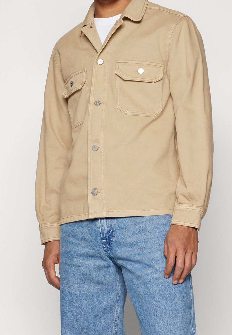 Ljust beige bomulljacka med två bröstfickor, knäppning med knappar och långa ärmar; kombinerad med ljusblå jeans.