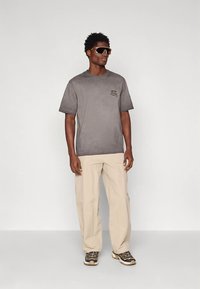 Muškarac nosi preveliku sivu majicu, bež hlače, smeđe i bež tenisice, te velike sunčane naočale smešnog tona, stoji ispred jednostavne bijele pozadine.