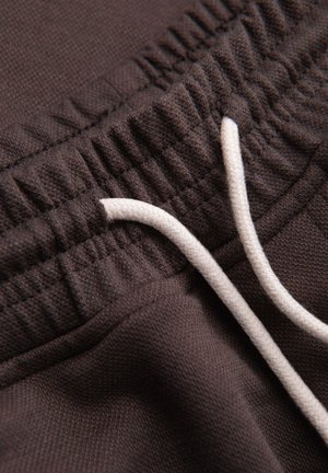 Close-up van een donkerbruine elastische tailleband met wit trekkoord op een kledingstuk van gestructureerde stof.