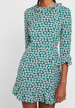 Frau trägt ein grünes Kleid mit weißem und pinkfarbenem Blumenmuster, dreiviertel Ärmeln und einem Rüschensaum sowie Rüschenmanschetten.