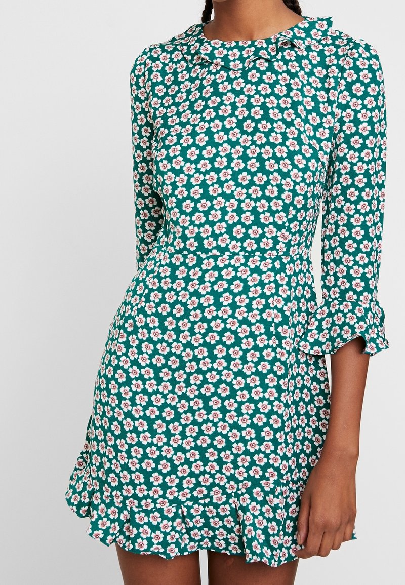 Femme portant une robe verte avec un imprimé de fleurs blanches et roses, manches trois-quarts, ourlet et poignets à volants.