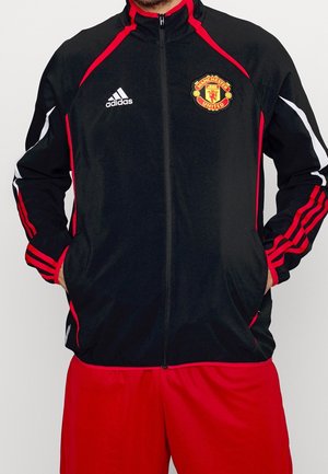 Person trägt eine schwarz-rote Manchester United Adidas Zip-Jacke mit den Händen in den Taschen und rote Sportshorts.