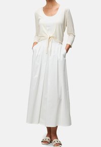 Bílý maxi bavlněný šaty se záhyby a bočními kapsami, doplněné krémovým kardiganem svázaným v pase. Oblečení je doplněno volnočasovými sandály a tříčtvrtečními rukávy.