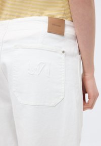 Άτομο που φοράει λευκό τζιν με δερμάτινη ετικέτα Mos Mosh και κεντημένο λογότυπο στην πίσω τσέπη, συνδυασμένο με πουκάμισο με κίτρινες ρίγες.