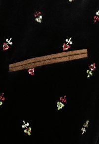 Tissu noir avec de petits motifs floraux brodés en rouge, blanc et vert, et un détail cousu horizontal marron près du centre.
