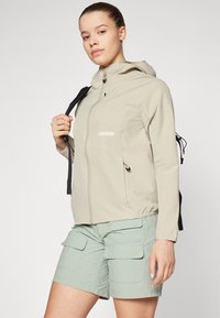 Veste zippée beige avec capuche, dotée d'une poche avant et d'un cordon de serrage, associée à un short cargo vert clair avec poches à rabat.