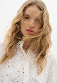 Camicia bianca con bottoni, caratterizzata da un motivo a griglia con ritagli. Il tessuto è leggero, texturizzato e semi-trasparente, con colletto e dettagli dei bottoni.