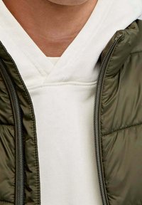 Grüne gesteppten Pufferjacke, die über einem weißen Hoodie mit V-Ausschnitt geöffnet ist. Texturierte Stoffdetails sind auf beiden Kleidungsstücken sichtbar.