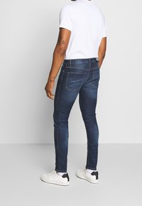 Tmavě modré denimové džíny s úzkým střihem, opatřené kontrastním šitím, pěti kapsami a označeným pasem. Nosené s jednoduchým bílým tričkem a teniskami.