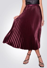 Gonna plissettata bordeaux realizzata in tessuto lucido, con vita alta e una linea fluida, abbinata a sandali neri con cinturini.