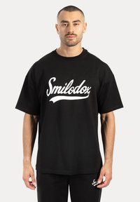 Svart bomulls-t-shirt med kort ärm, med en stor vit "Smilodox"-logotyp över bröstet. Avslappnad passform med rund halsringning.