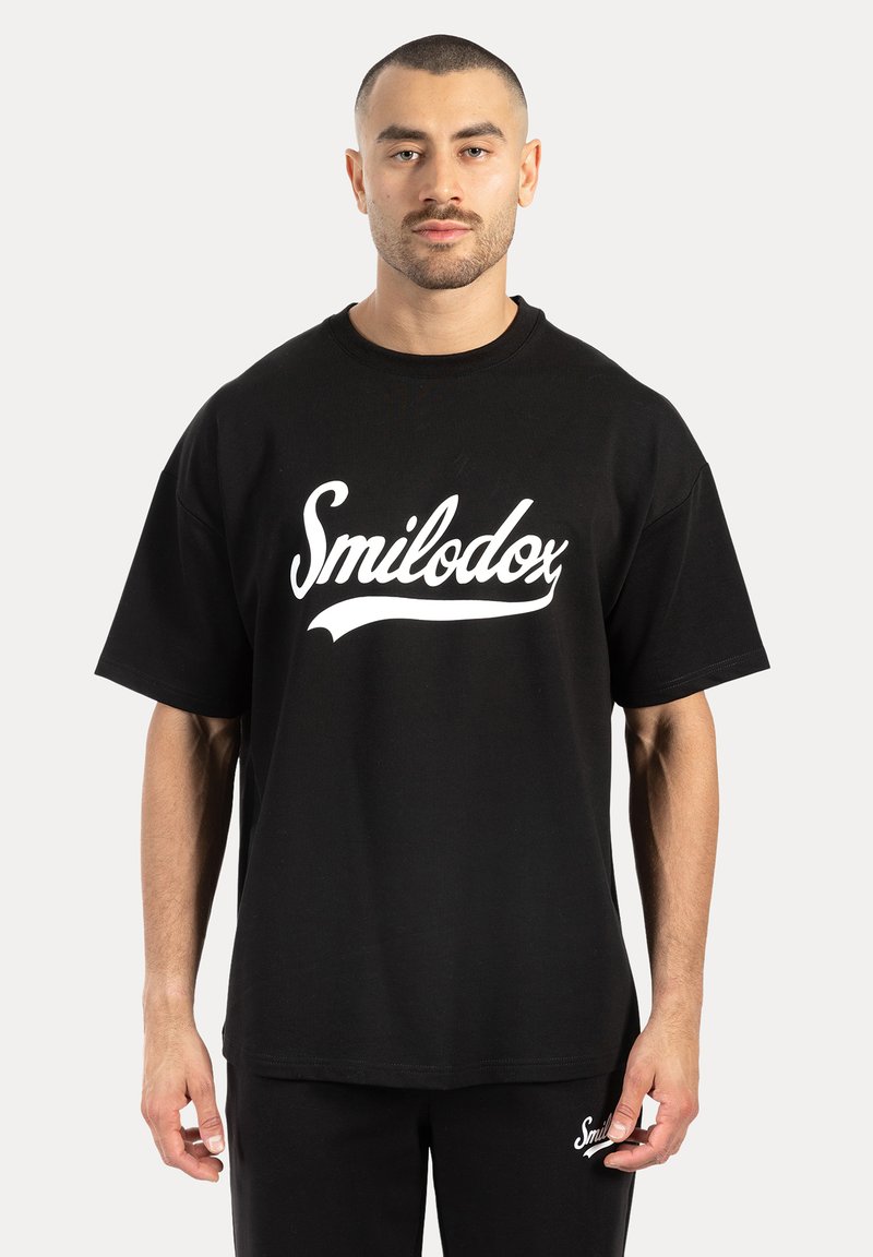 Svart bomulls-t-shirt med kort ärm, med en stor vit "Smilodox"-logotyp över bröstet. Avslappnad passform med rund halsringning.