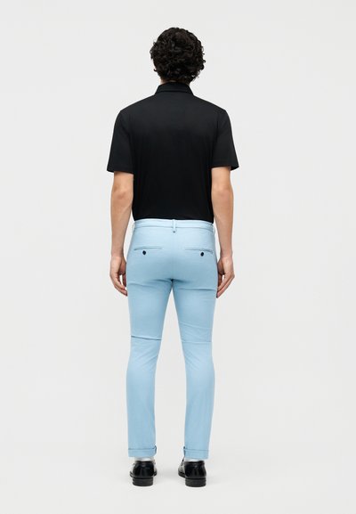 Άνδρας με σγουρά μαλλιά που φοράει μαύρο κοντομάνικο πουκάμισο, ανοιχτόχρωμο μπλε παντελόνι στενής γραμμής και μαύρα παπούτσια, στέκεται με την πλάτη γυρισμένη.