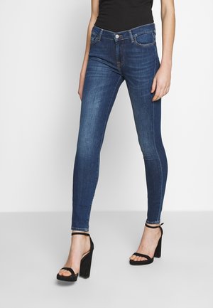 Slanke mørkeblå jeans med sorte højhælede sandaler og en sort top stukket ind, vist fra midt på låret og ned mod en ensfarvet baggrund.